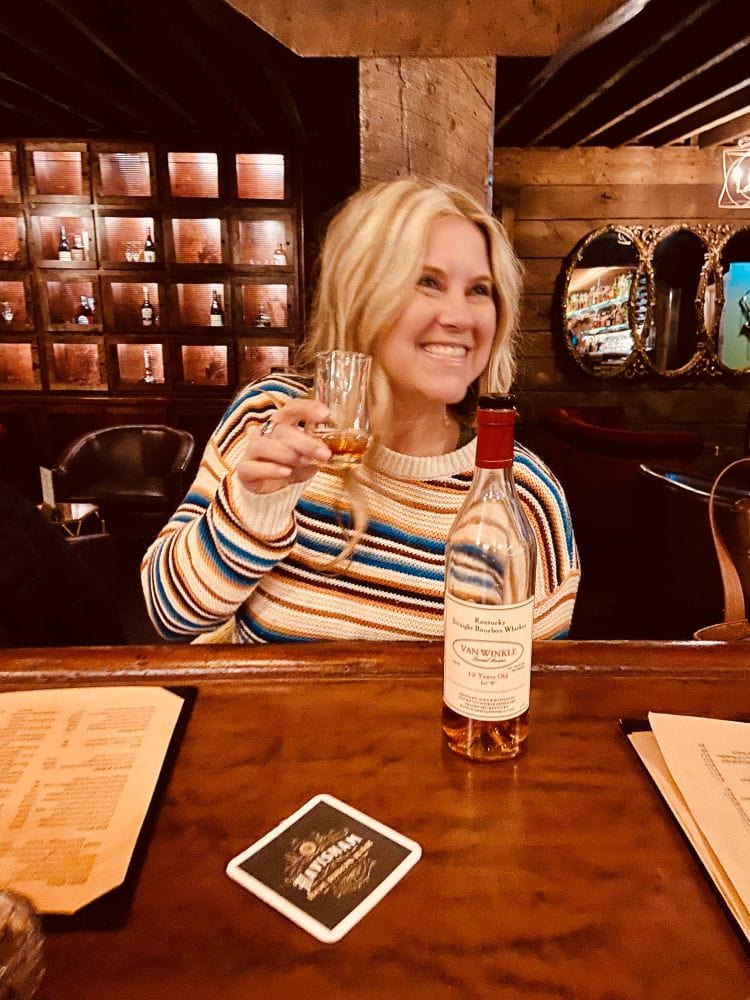 Me holding up a glass of bourbon, a bottle of Pappy's in front of me, at the bar at Havisham Bourbon Bar.