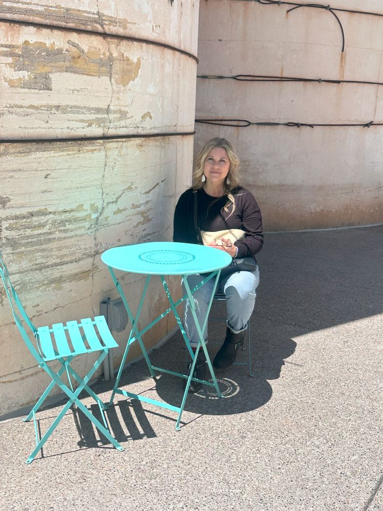 Bev is sitting outdoors at a turquoise bistro chair in front of the white silos at Los Poblanos, an Albuquerque lavender farm.