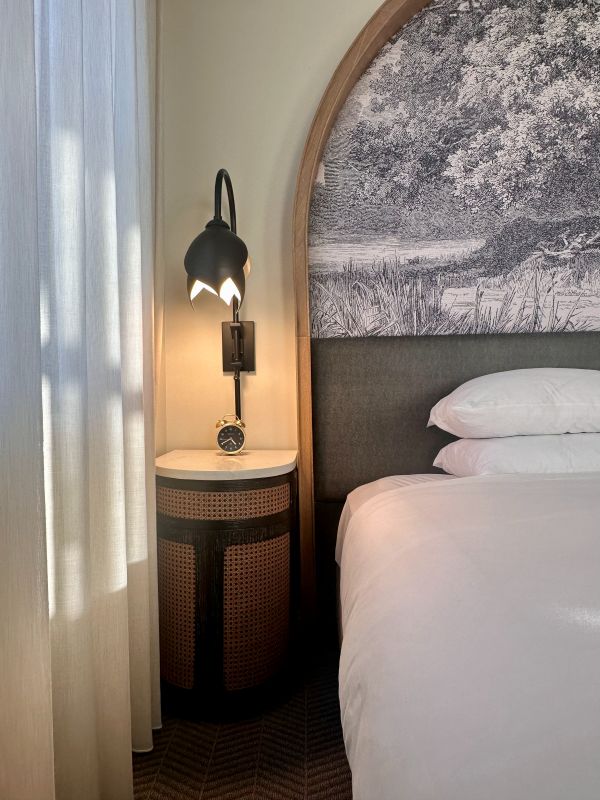 A partial view of the bed and fabric headboard and side table with lamp in a guest room at The Cottonwood Hotel, a cozy holiday stay in Omaha.