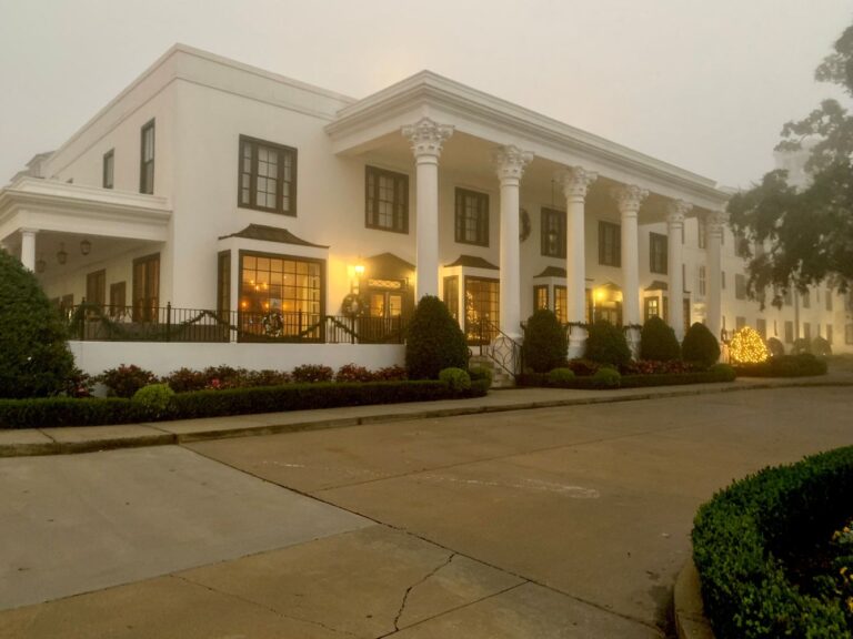 The exterior of the White House Hotel, a white structure with columns, in the fog with yellow light illuminating the front, a Gulf coast getaway.