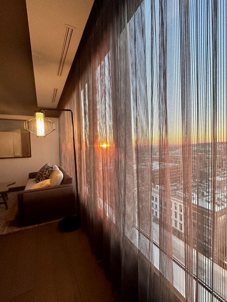A guest room with a wall of windows, sheer curtains and couch, the sunset seen through the curtains at The Farnam, one of the best hotels in Omaha.
