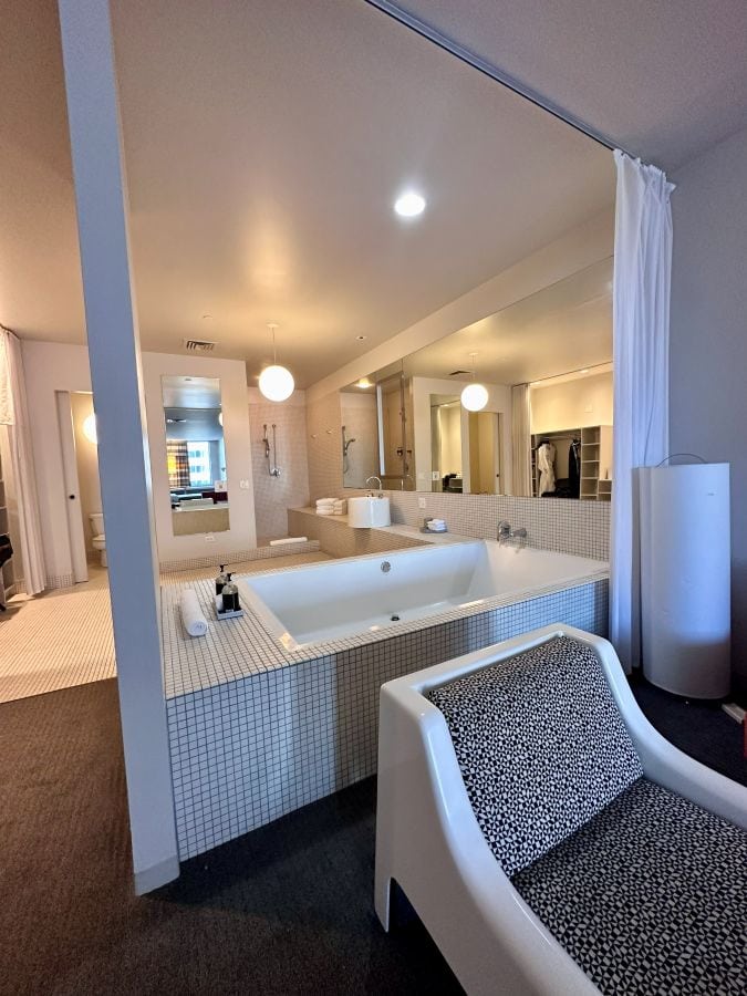 A modern white plastic like chair with cushions in the foreground, a huge soaking tub and bathroom in the background in a room at The Delphi, a boutique hotel downtown Los Angeles.