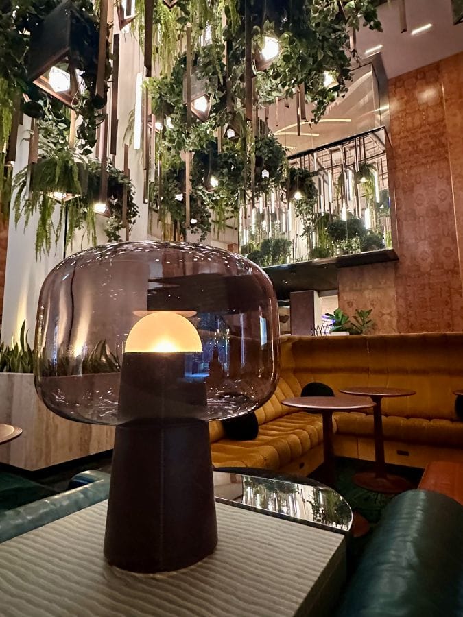 A modern gray domed art deco lamp on a table with greenery from the ceiling in the background and warm wood walls in the lobby of The Dephi, a boutique hotel downtown Los Angeles.