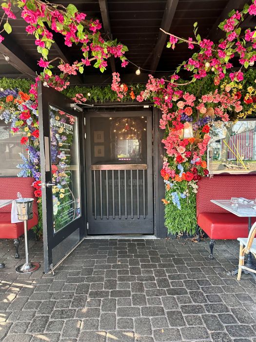 The entrance under the patio rood with a swing door and pink flowers surrounding it at Petite Chou Bistro and Champagne Bar a local restaurant for breakfast in Indianapolis.