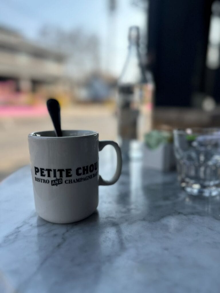 A white mug of coffee on the outdoor table with a home in the background at Petite Chou Bistro and Champagne Bar, a local restaurant for breakfast in Indianapolis.