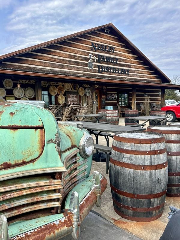 Exterior of Neeley Family Distillery, old Chrysler and bourbon barrels in front.
