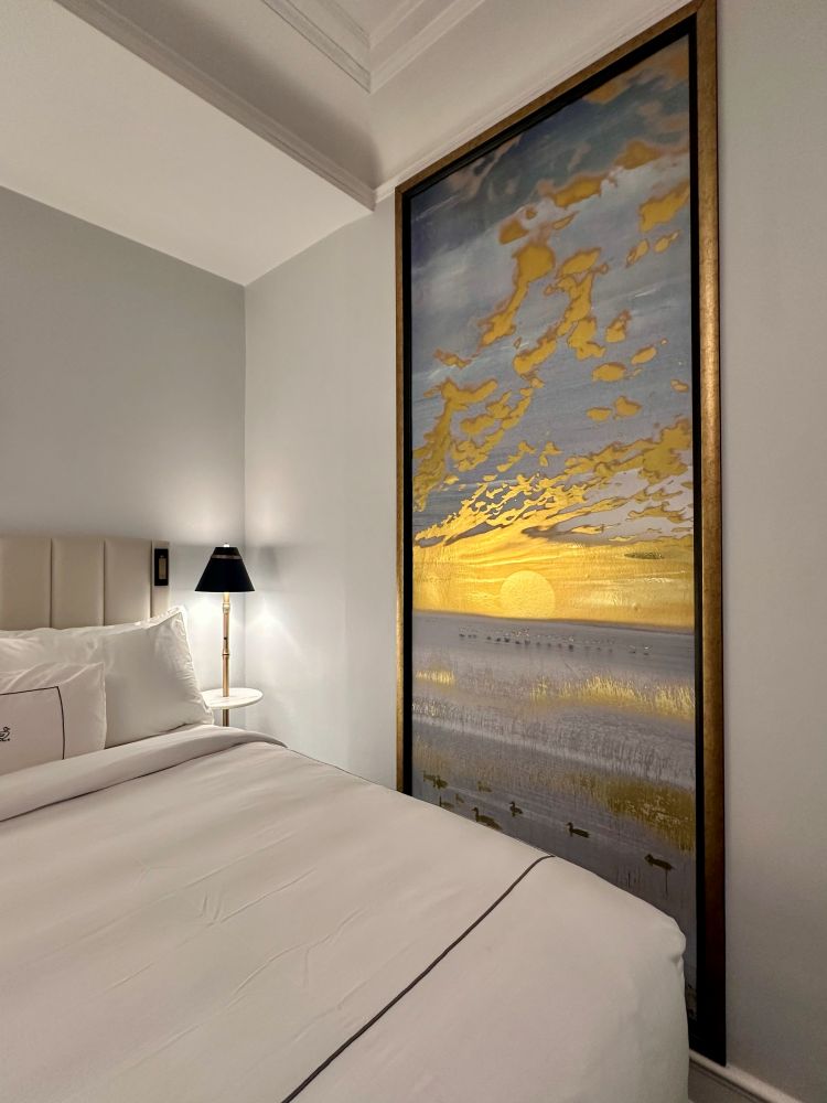 A guest bedroom, the guest bed in white, pale gray walls and a gold and blue large artwork on the wall at the Peabody Hotel in Memphis TN.