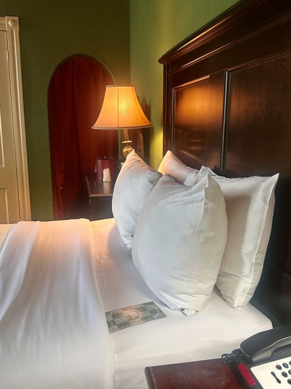 Pillows on a bed with a dark wood headbpard, warm light table lamp in the background, green walls, a guest room at the 1886 Crescent Hotel Eureka Springs.