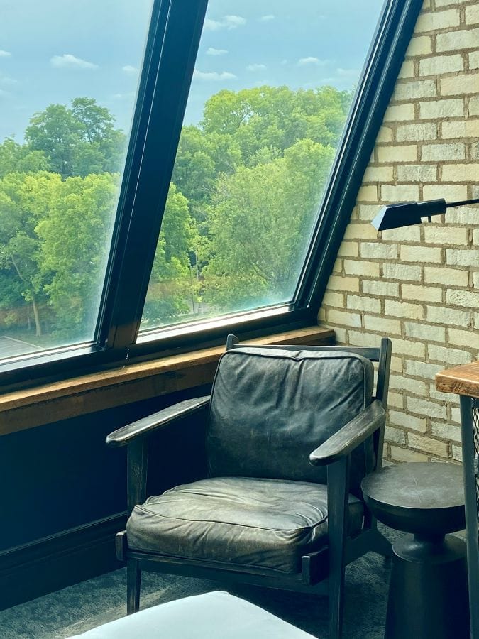 A dark leather and wood chair in the corner of a guest room next to the large angled windows in a guest room at the Ironworks Hotel, an industrial style hotel in Indianapolis.