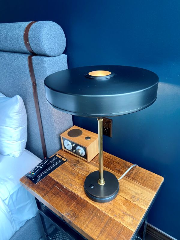 A unique disc-style deco lamp in dark blue, dark blue walls, and a side table next to the bed in the guest room at the Ironworks Hotel, an industrial-style hotel in Indianapolis.