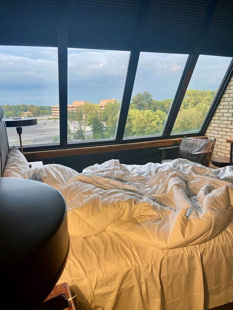 A guest room, a messy bed in the foreground and a wall of large windows with a moody sky in the background at the Ironworks Hotel, an industrial style hotel in Indianapolis.