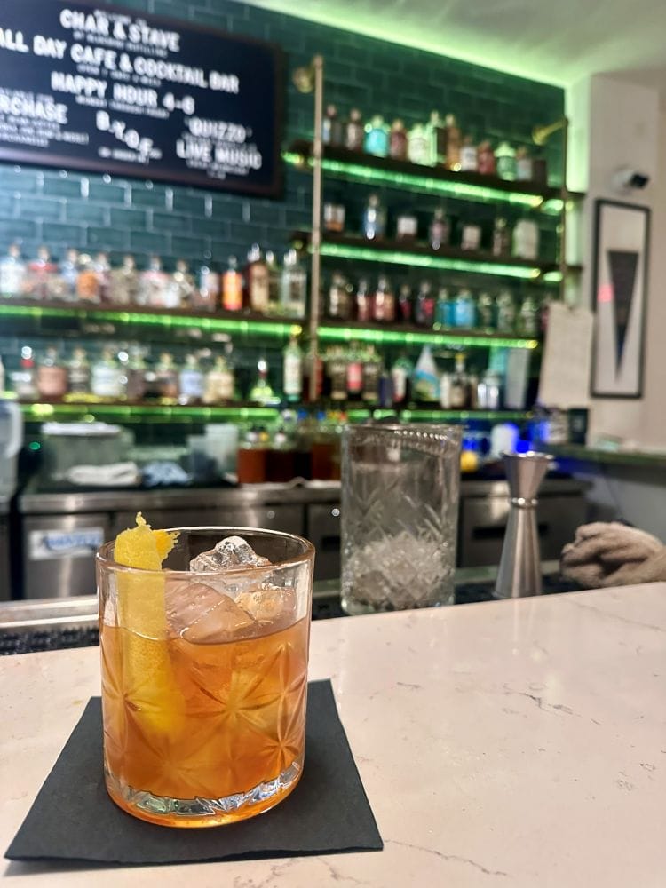 An Old Fashioned cocktail on the bar with the bar of bottles in the background at Char & Stave, a Cocktail and Coffee bar in Chestbut Hill, Philadelphia off the beaten path.