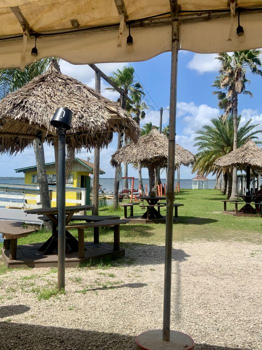 Straw umbrellas over round wood tables with a bue sky and the lake in the background at Lazy Gator Bar.