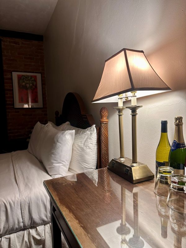 A side view of pillows on a guest bed and a side table and lamp, with bottles of wine on the table that can be purchased to use on the balcony at the Park Plaza Hotel, a Winter Park Florida hotel.