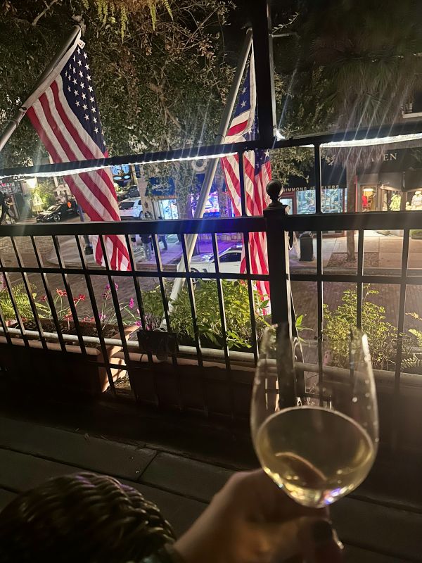 A glass of wine on the table, the iron banister in the background with the American Flags hanging at night, lights shinning on them on the balcony at the Park Plaza Hotel, a Winter Park Floriday Hotel.