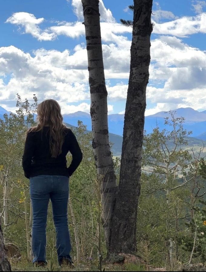 Bev back to the camera standing next to a tree with the mountain range in the background on my Colorado mountain retreat.