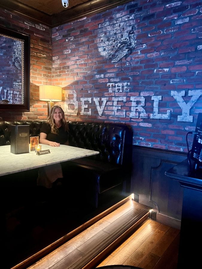 Cozy bar interior with brick walls and ambient lighting in Scottsdale. Bev sitting in front of a painted sign on the brick wall "The Beverly".